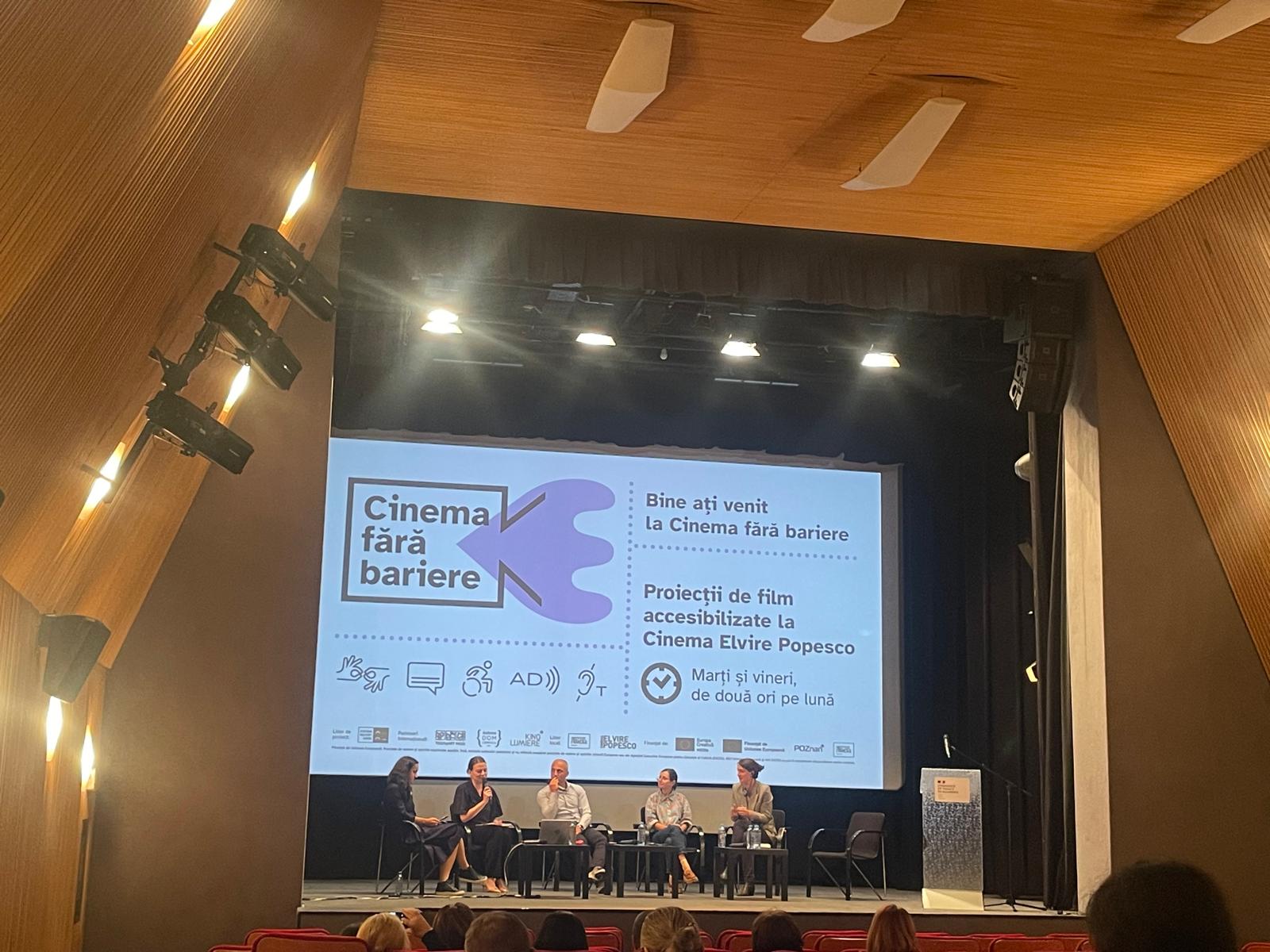 Photo taken from the cinema hall. The stage is visible and there 5 speakers sitting on chairs.